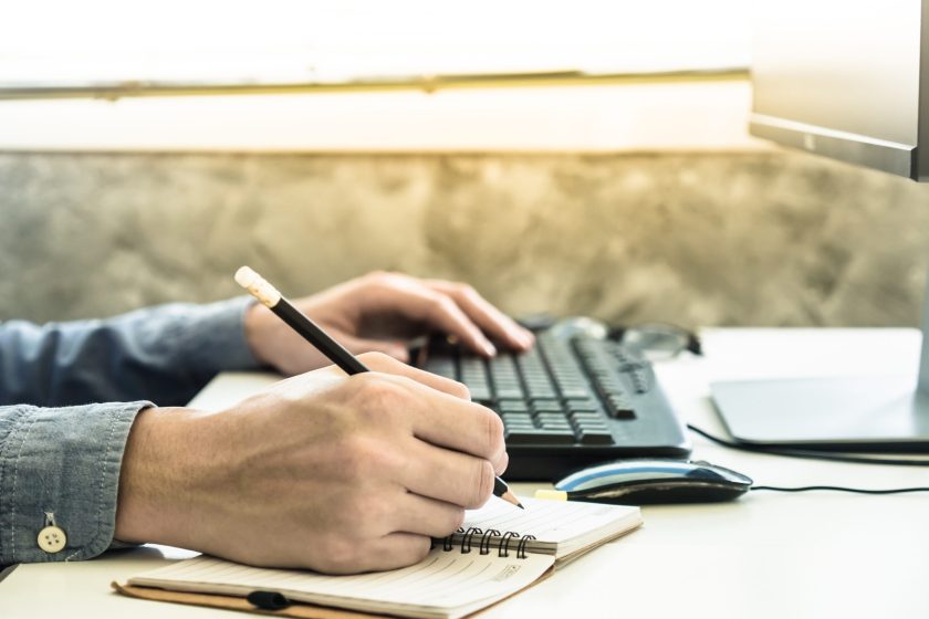 Close up of Male hands writing some data in notebook and using computer on the desk